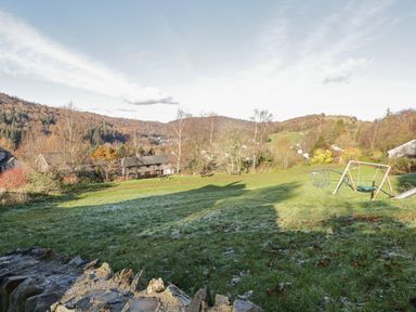 A grassy open area with a stone wall in the foreground playground equipment and houses with hills in the background at Bishop Fell in Newby Bridge