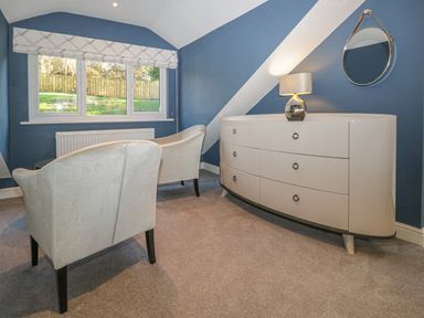 A room with two chairs facing a window and a curved dresser with a lamp and round mirror on a blue wall at Bishop Fell in Newby Bridge