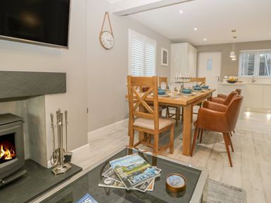 A dining area with a wooden table set with dishes and glasses next to a fireplace and a glass coffee table with magazines in an open plan kitchen dining space at Bishop Fell in Newby Bridge