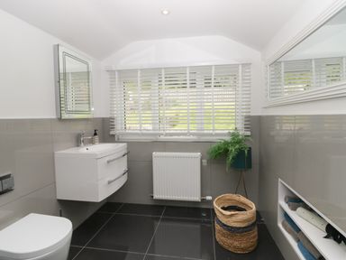 A bathroom with a white floating sink cabinet a toilet a window with white blinds a radiator a plant in a green pot and a wicker basket at Bishop Fell in Newby Bridge