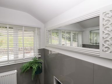 A room with a window covered by white blinds a decorative mirror a plant in a stand and a radiator at Bishop Fell in Newby Bridge