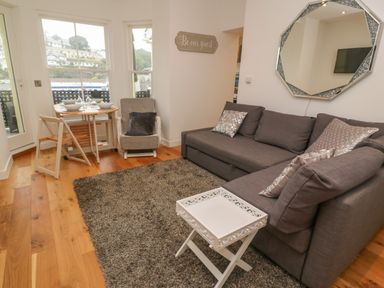 A living room with a gray sofa, a white tray table, a round dining table with chairs near windows at 1 River View in Looe