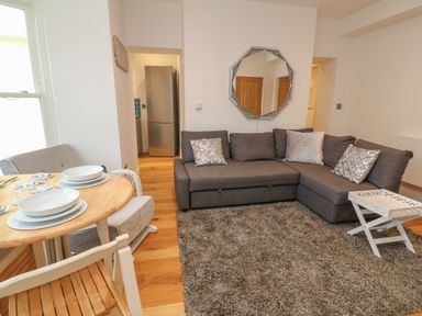 A living room with a gray corner sofa a round wooden dining table with place settings and a refrigerator visible in the doorway at 1 River View in Looe