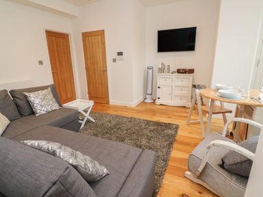 A living room with a gray sofa and silver pillows two wooden doors a white cabinet with a TV mounted above a small dining table with chairs at 1 River View in Looe