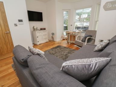 A living room with gray sofa and armchair a small dining table in front of windows and a white cabinet with a wall-mounted TV at 1 River View in Looe