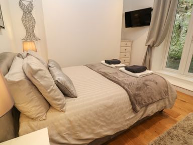A bedroom with a bed made with multiple pillows and folded towels on a blanket a wall-mounted TV and a window with curtains at 1 River View in Looe