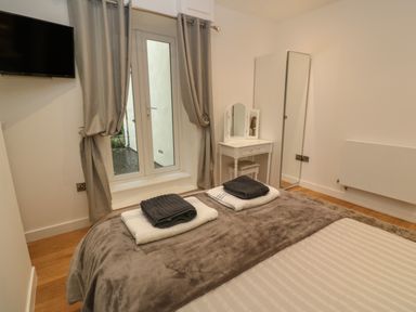 A bedroom with a bed, towels on the bed, a wall-mounted television, curtains, a small vanity table with mirror, and a tall freestanding mirror at 1 River View in Looe