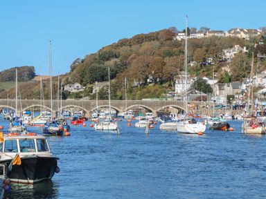 A river filled with moored boats and a stone arched bridge with houses on a hillside in the background at 1 River View in Looe