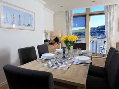 A dining room with a wooden table set for six with flowers on the table and a view of water and hills through windows at 2 Embankment House in Dartmouth