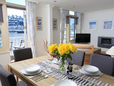A dining table set with plates glasses and cutlery with yellow flowers in a vase in a living room with a TV and fireplace at 2 Embankment House in Dartmouth