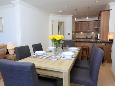 A dining area with a wooden table set with dishes and glasses and yellow flowers in a vase near a kitchen with wooden cabinets at 2 Embankment House in Dartmouth