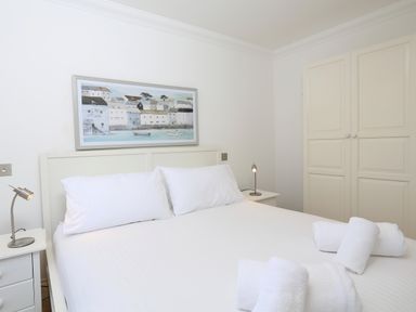 A bedroom with a white bed, two pillows, rolled white towels, nightstands with lamps, a wardrobe, and a framed painting above the bed at 2 Embankment House in Dartmouth
