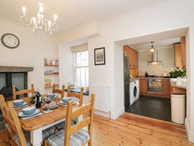 A dining area with wooden table and chairs set for a meal next to a kitchen with wooden cabinets and appliances at The Postmasters House in Windermere