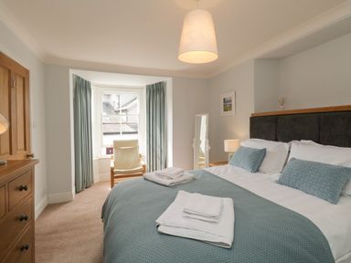 A bedroom with a bed blue pillows blue blanket and folded white towels a wooden chest of drawers and a chair by a window at The Postmasters House in Windermere