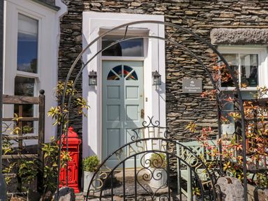 The entrance door of a stone house with a metal arch and gate with the words The Masters House a red mailbox and plants at The Postmasters House Windermere