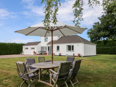 A garden with a wooden table and chairs under a large umbrella in front of a white house with a double garage at Trevore Farmhouse in Chacewater