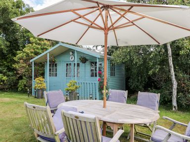 A garden with wooden table and chairs under an umbrella near a blue garden shed at Trevore Farmhouse in Chacewater