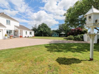 A white house with a paved driveway a green lawn and a wooden birdhouse on a post at Trevore Farmhouse in Chacewater