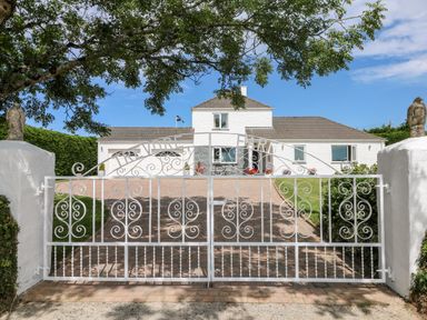 A white farmhouse behind a decorative white iron gate with trees and a paved driveway at Trevore Farmhouse in Chacewater