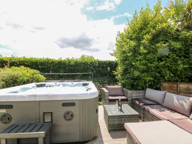 An outdoor patio with a hot tub a wicker sofa set and a glass table with a bottle and glasses at Trevore Farmhouse in Chacewater