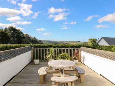 An outdoor wooden deck with a round picnic table and benches with potted plants and countryside views at Trevore Farmhouse in Chacewater