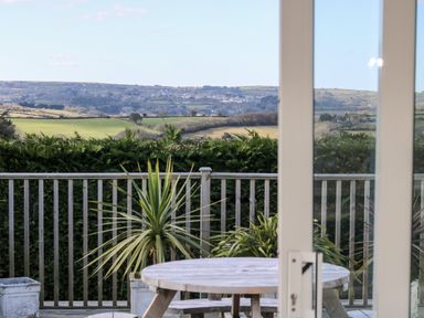 A wooden deck with a round table and potted plants overlooking green fields at Trevore Farmhouse in Chacewater