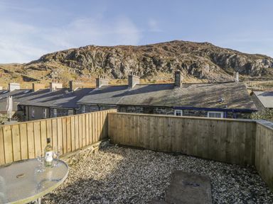 An outdoor patio area with a glass table holding a bottle and two glasses surrounded by a wooden fence and stone houses with hills in the background at Manod View in Blaenau Ffestiniog