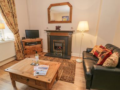 A living room with a black sofa with cushions a wooden coffee table with magazines a fireplace a wooden TV stand with a TV and a floor lamp at Manod View in Blaenau Ffestiniog