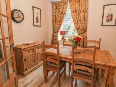 A dining room with a wooden table and chairs a wooden sideboard a window with curtains and flowers on the table at Manod View in Blaenau Ffestiniog