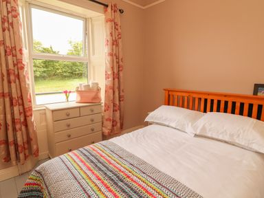 A bedroom with a wooden bed with white bedding and a colorful blanket next to a chest of drawers and a window with floral curtains at Newtown East in Newtown East near Kilkee County Clare