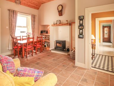 A dining area with wooden table and chairs near a window a wood stove under a wall clock and an armchair with colorful cushions at Newtown East near Kilkee County Clare