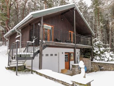 A snow-covered chalet with a balcony and stairs in a forested area at Torbreck Chalet in Kincraig