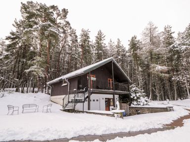 A cabin in a snowy wooded area with outdoor chairs and a pathway covered in snow at Torbreck Chalet in Kincraig