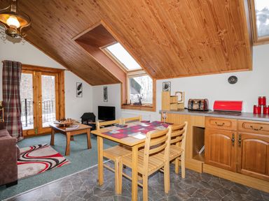 A dining area with a wooden table and chairs next to a kitchen countertop and a living area with a coffee table chess set and a television at Torbreck Chalet in Kincraig