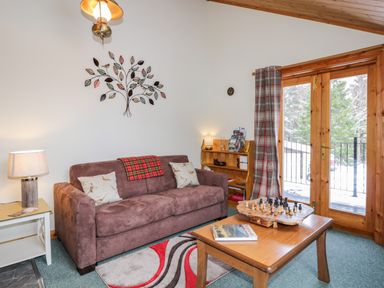 A living room with a brown sofa a wooden coffee table with a chess set and book a side table with a lamp and a wooden cabinet next to glass doors with plaid curtains at Torbreck Chalet in Kincraig