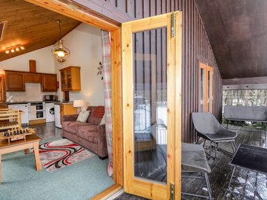 A kitchen and living area with wooden cabinets brown sofa and a rug next to an outdoor balcony with chairs and a table at Torbreck Chalet in Kincraig