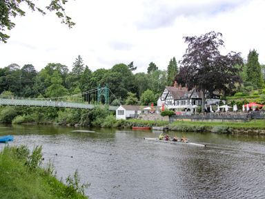 A river with rowers in a boat a suspension bridge and a pub with outdoor seating at The Boars Head Pub in Bishop's Castle