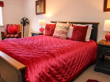 A bedroom with a red bedspread pillows two bedside tables lamps a striped armchair and a standing fan at The Boars Head Pub Bishop's Castle