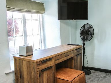 A wooden desk with a tissue box on top and a brown ottoman underneath next to a window with a wall-mounted TV and a standing fan at The Boars Head Pub in Bishop's Castle