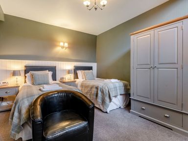 A bedroom with two single beds, bedside tables with lamps, a black armchair, and a wardrobe at The Boars Head Pub in Bishop's Castle