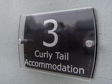 A black and clear rectangular sign with the number 3 and the words Curly Tail Accommodation on a gray wall at The Boars Head Pub in Bishops Castle