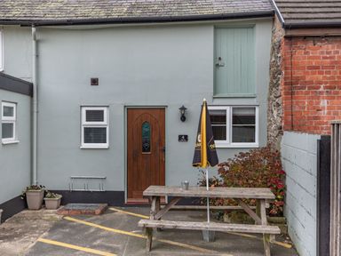 An outdoor area with a wooden bench and table with an umbrella next to a grey building with a wooden door and windows at The Boars Head Pub in Bishop's Castle