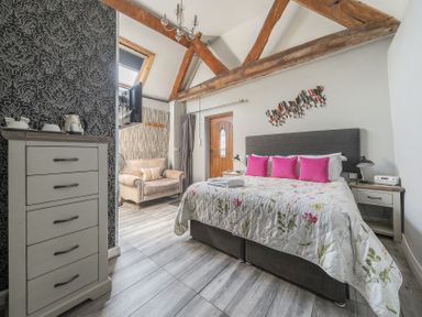 A bedroom with a large bed and pink pillows a dresser and an armchair with exposed wooden beams at The Boars Head Pub Bishop's Castle