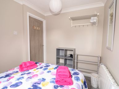 A bedroom with a bed with colorful bedding and pink towels a cubby shelf with a hairdryer a clothes rack a door and a mirror on the wall at Little Wash Cottage in Hunstanton