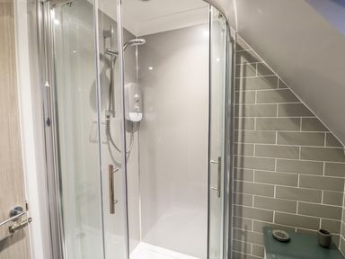 A bathroom with a glass shower enclosure a wall-mounted shower unit and grey tiled walls at Little Wash Cottage in Hunstanton