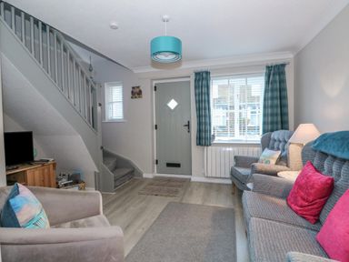 A living room with a gray sofa with pink cushions an armchair a wooden TV stand with a television stairs with gray railing and a front door with a diamond window at Little Wash Cottage in Hunstanton