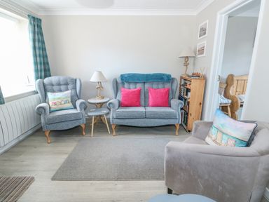 A living room with armchairs a sofa side tables lamps a bookshelf and pink cushions at Little Wash Cottage in Hunstanton