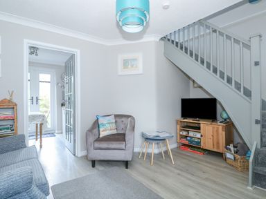 A living room with a gray armchair a wooden TV stand under a staircase and a doorway leading to a dining area at Little Wash Cottage in Hunstanton