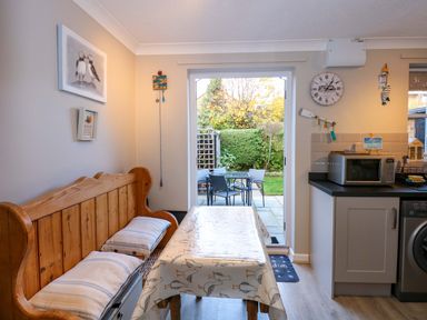A dining area with a wooden bench and table covered with a patterned tablecloth next to a kitchen counter with a microwave and washing machine and a glass door leading to an outdoor patio with chairs and a table at Little Wash Cottage in Hunstanton