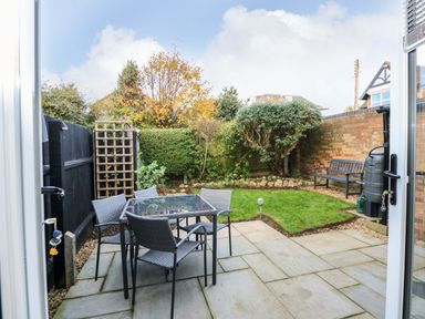 A backyard patio with a glass table and four chairs a grassy area with plants and a bench at Little Wash Cottage in Hunstanton
